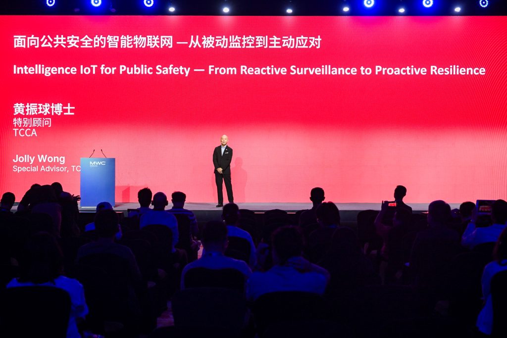 A speaker in a suit stands onstage before a seated audience. A large red screen behind him displays bilingual text: “Intelligence IoT for Public Safety — From Reactive Surveillance to Proactive Resilience” and his name, Jolly Wong.