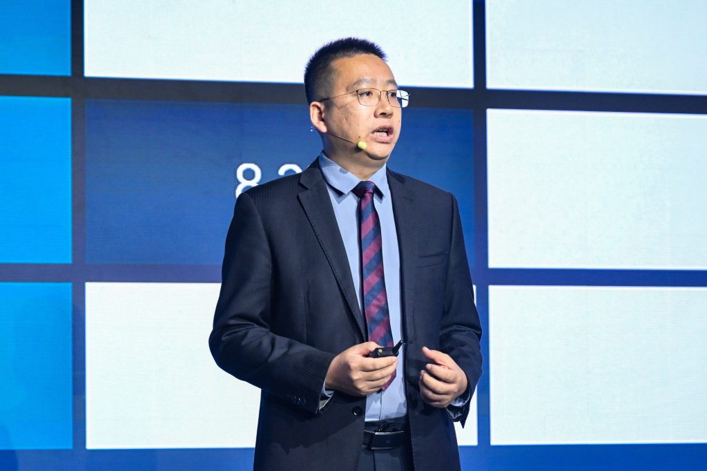 A man in a suit and striped tie speaks onstage, wearing glasses and a headset microphone. He holds a small device and stands before a blue and white geometric background with numbers partially visible on the screen.