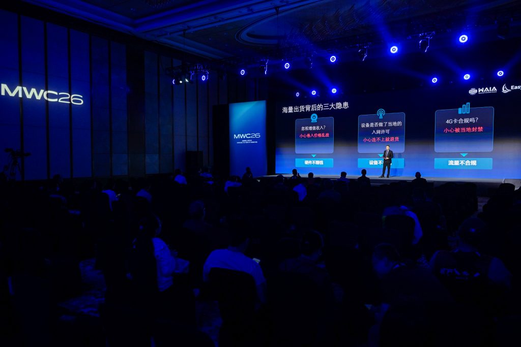 A speaker stands on a stage at a tech conference, "MWC26," in a dark auditorium. Blue lighting highlights large screens behind the speaker displaying Chinese text and graphics. Audience members sit in rows, mostly in silhouette, facing the stage.