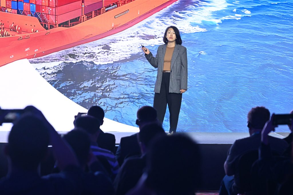 A woman in a gray blazer and brown top stands on stage, giving a presentation. Behind her is a large image of a cargo ship at sea. Audience members are visible in silhouette, some holding up phones to record or take photos of the presentation.