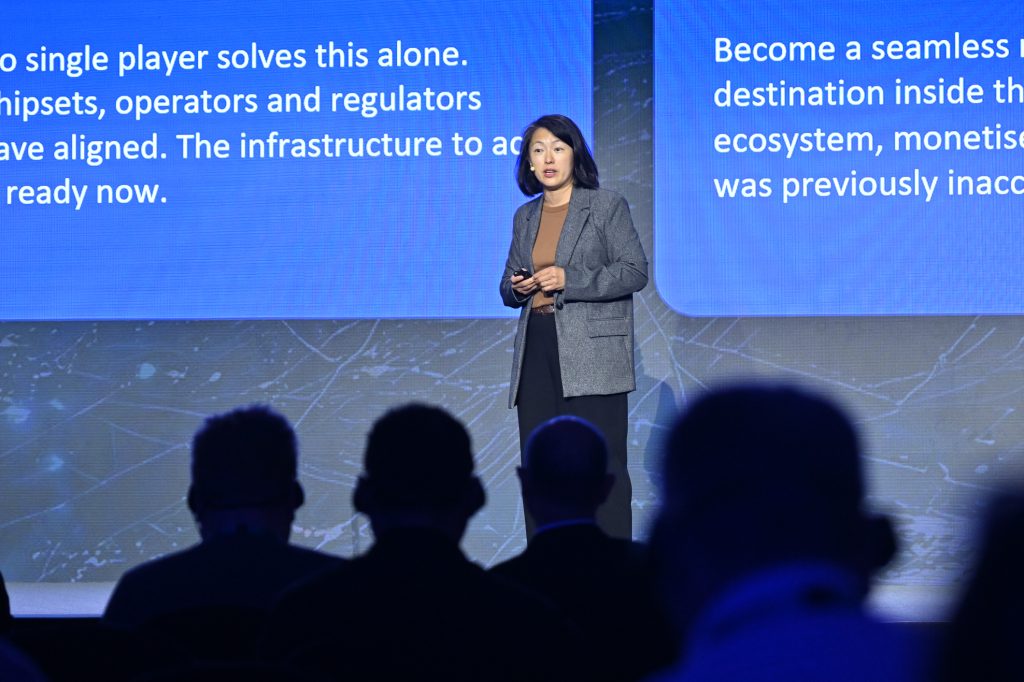 A woman in a gray blazer speaks on stage at a conference. Blue screens behind her display white text. Several people are seated in the dark foreground, watching her presentation. The setting appears professional and focused on technology or business.