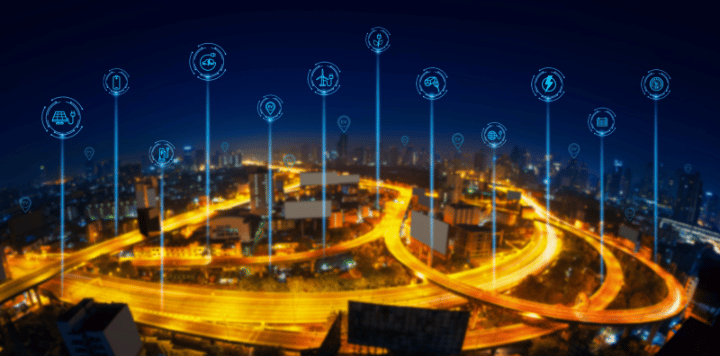 A cityscape at night shows glowing highways and buildings, with blue digital icons above representing smart city technologies like energy, transportation, and communication, interconnected across the skyline.