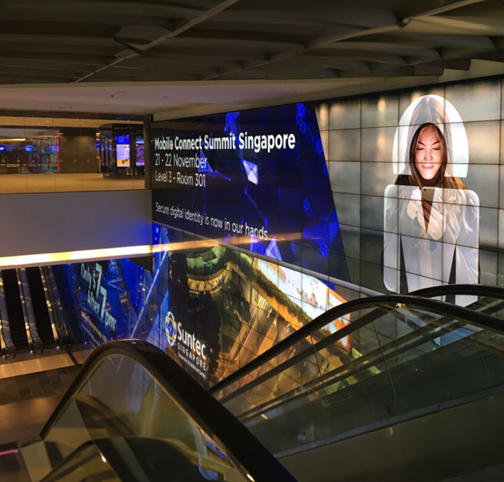 An illuminated billboard advertising the mobile connect summit in singapore, overlooking an escalator inside a modern building.