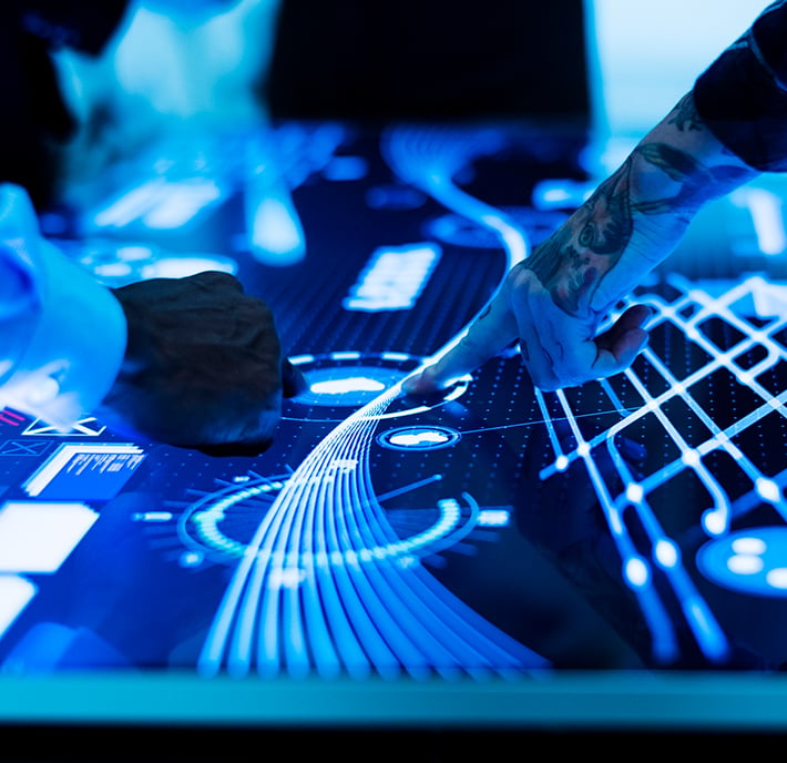 Close-up of multiple hands interacting with a large, touchscreen display emitting blue light. The display shows various digital graphics and data visualizations. One hand has visible tattoos, and people are standing around the table.