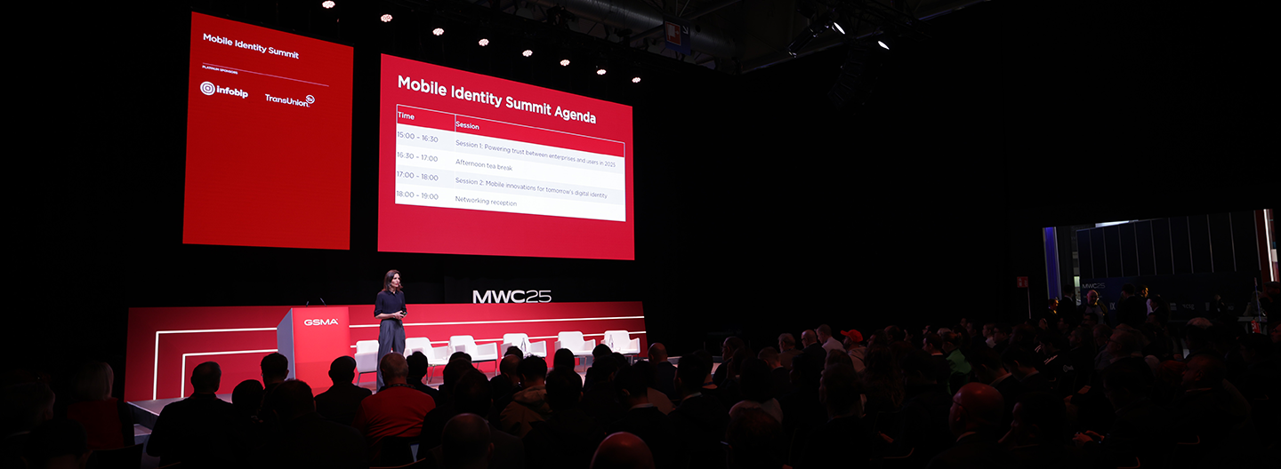 A presenter takes the stage at the Mobile Identity Summit, where identity and data are in focus. Behind them, a large screen shows the agenda against a red background with logos. The audience sits attentively in the dimly lit venue, spotlighting highlighting the speaker.