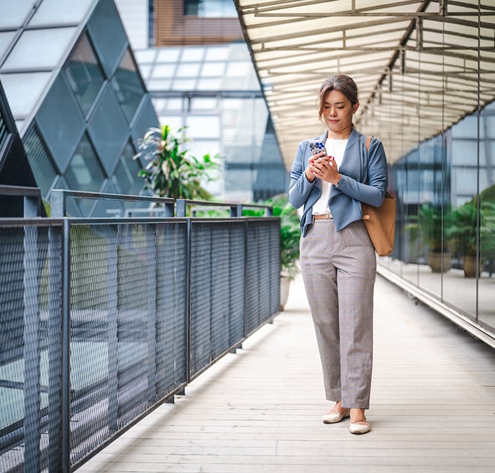 A woman in business attire walks on an outdoor walkway, checking her phone for identity and data updates. She wears a blue blazer, white top, grey checked trousers, and beige flats, carrying a tan bag amid glass buildings and greenery in a modern urban setting.