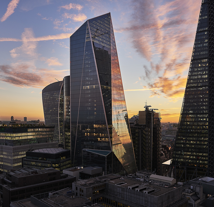Tall, modern skyscrapers with glass façades reflect a colourful sunset sky streaked with clouds, symbolising the city’s evolving identity and data-driven growth. The angular buildings dominate the cityscape, glowing warmly on the horizon.