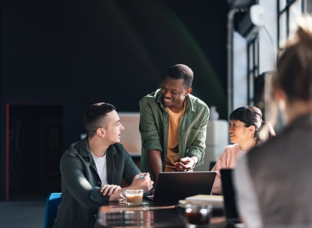 Three people sit around a table with laptops and coffee in a sunlit modern office, discussing how to number verify availability. One man stands, smiling as he talks to the attentive group, whilst another person is blurred in the foreground.