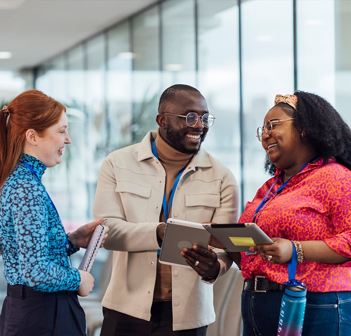 Three colleagues stand together in a bright office space, smiling and talking about identity and data. One holds a notebook, another a tablet, and the third a clipboard. Large windows are in the background as they collaborate on their project.