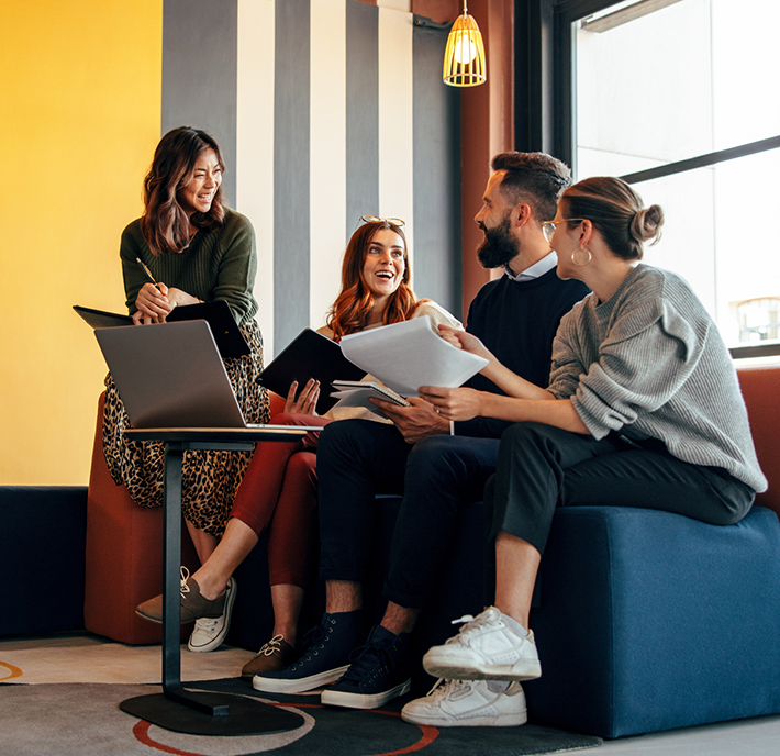 Four young adults sit together on modern seating in a stylish, colourful room with a laptop and papers, smiling and engaging in conversation about identity and data. Natural light comes through a large window as they collaborate or study.