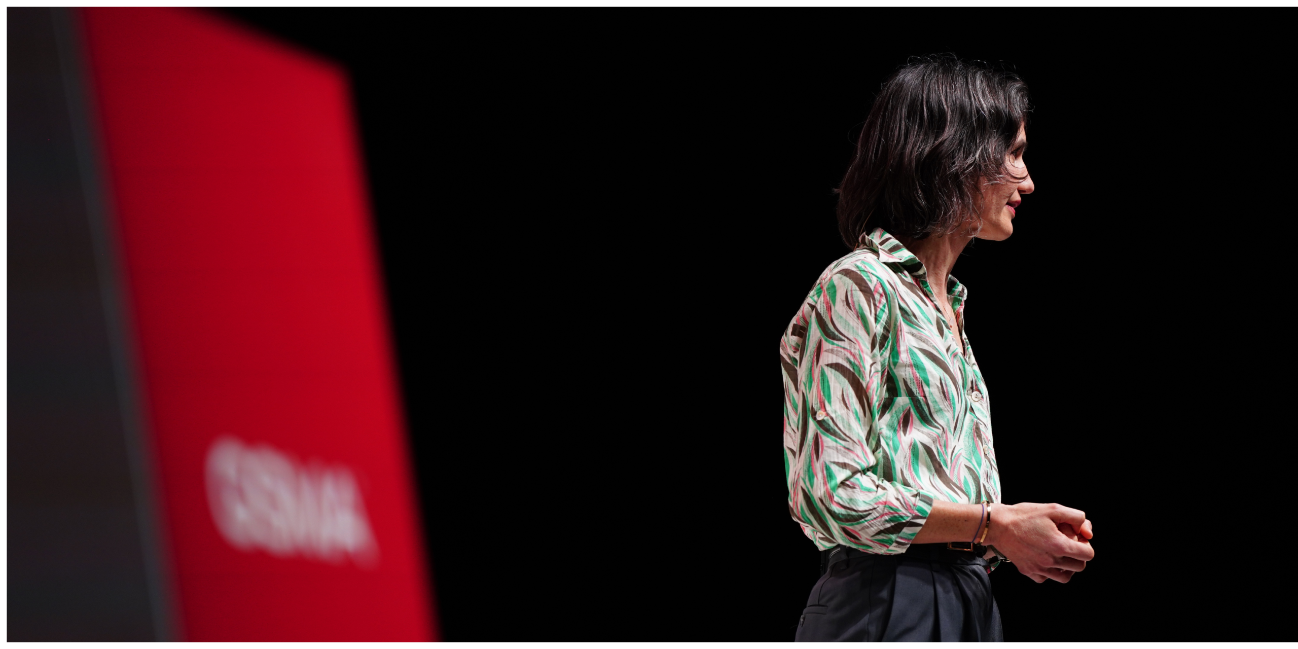 Person with shoulder-length dark hair gives a talk on stage, standing near a red screen with blurred text.