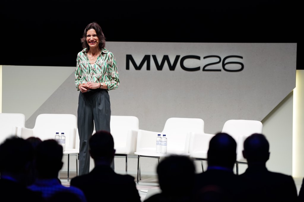 A woman speaks on stage in front of an "MWC26" sign, with empty chairs and an audience visible.