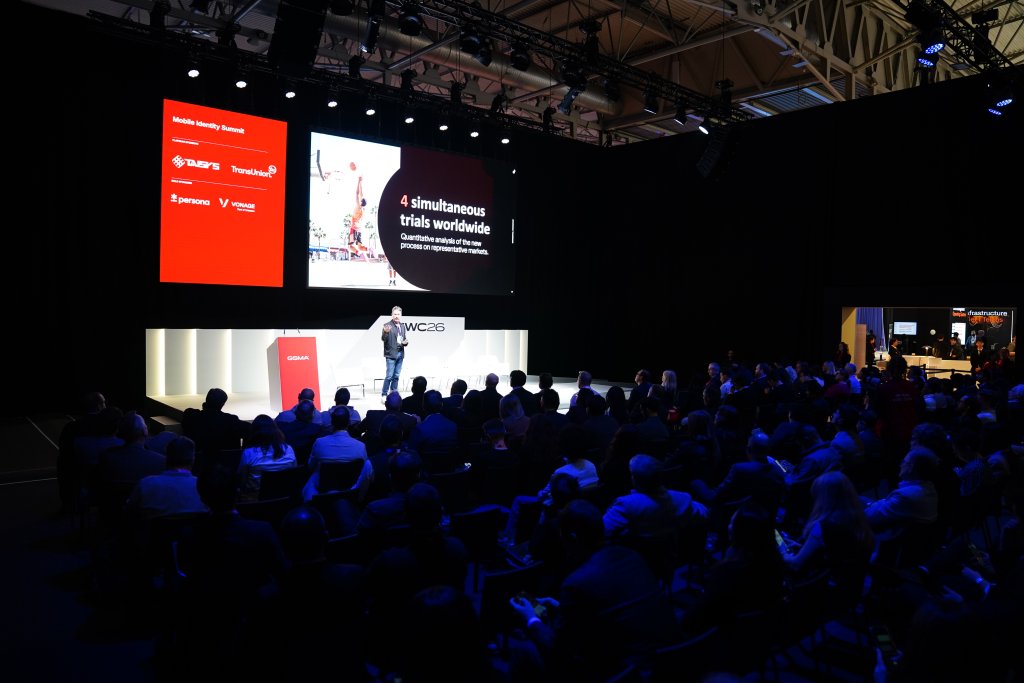 A speaker presents on stage to an audience at a tech conference, large screens display event information.