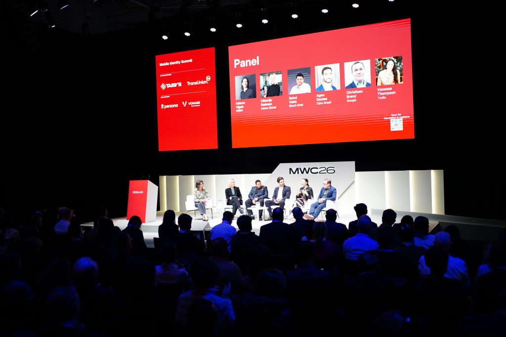 A panel of six speakers discusses on stage at MWC24, with a large red screen displaying panel info behind them.