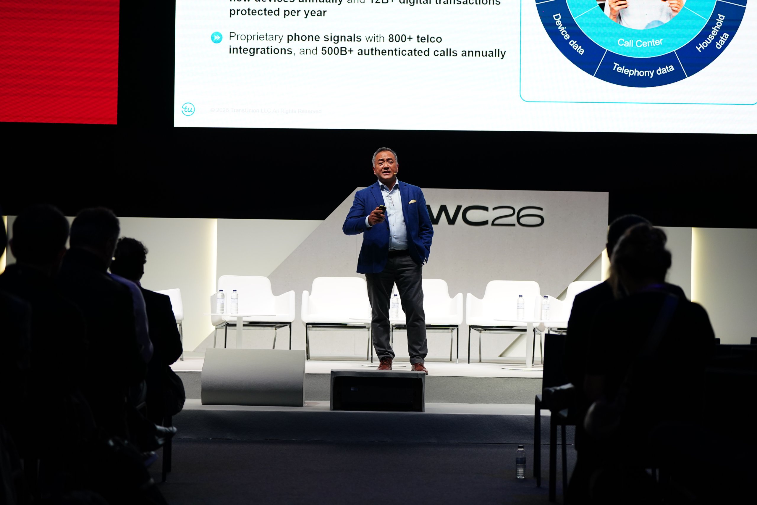 A man in a blue blazer speaks on stage at a conference, with a presentation slide projected behind him.