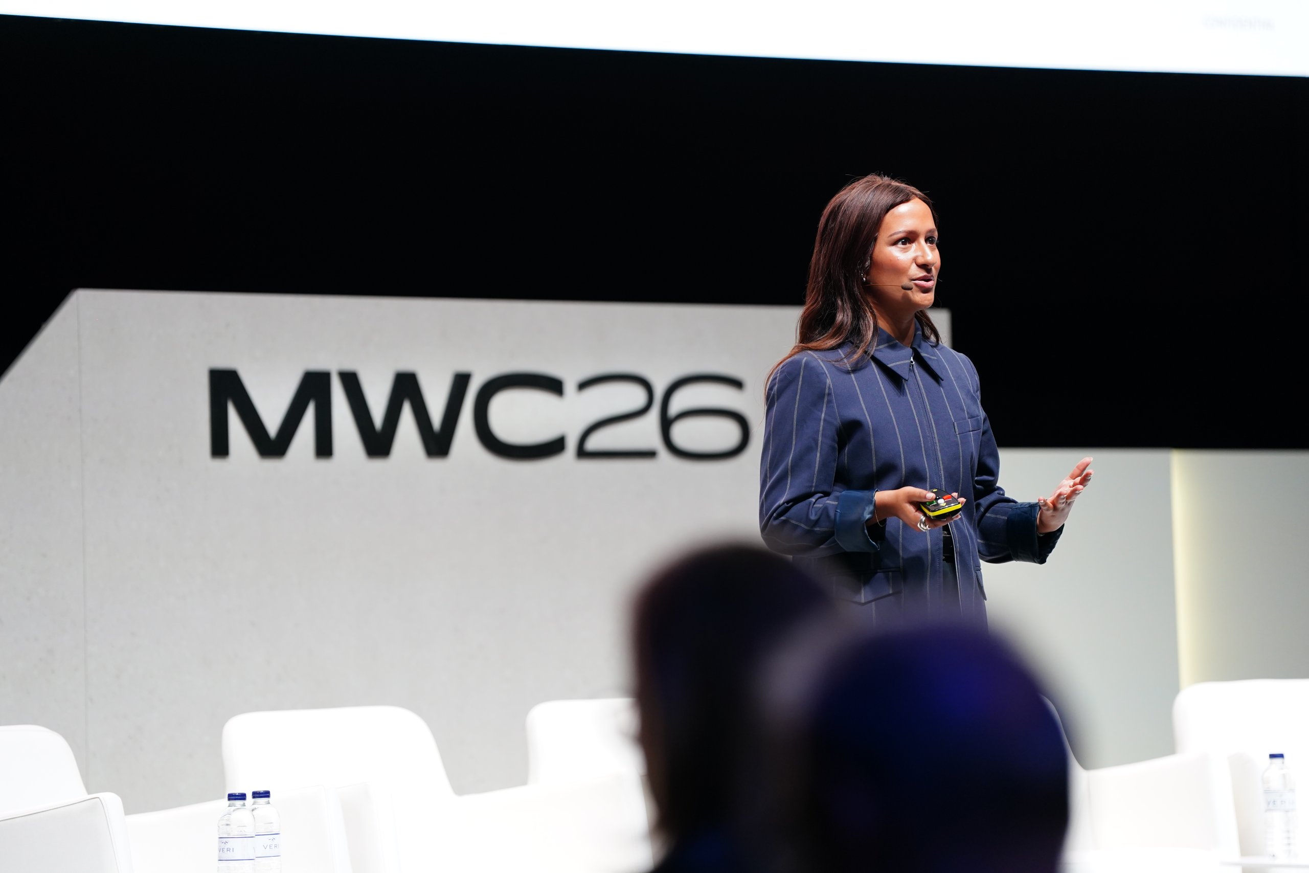 A woman speaks on stage at MWC 26, holding a device, with an audience in the foreground.