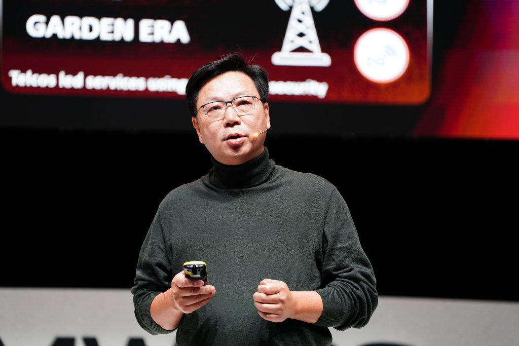 Man in glasses and dark sweater speaks on stage holding a device, with presentation screen behind him.