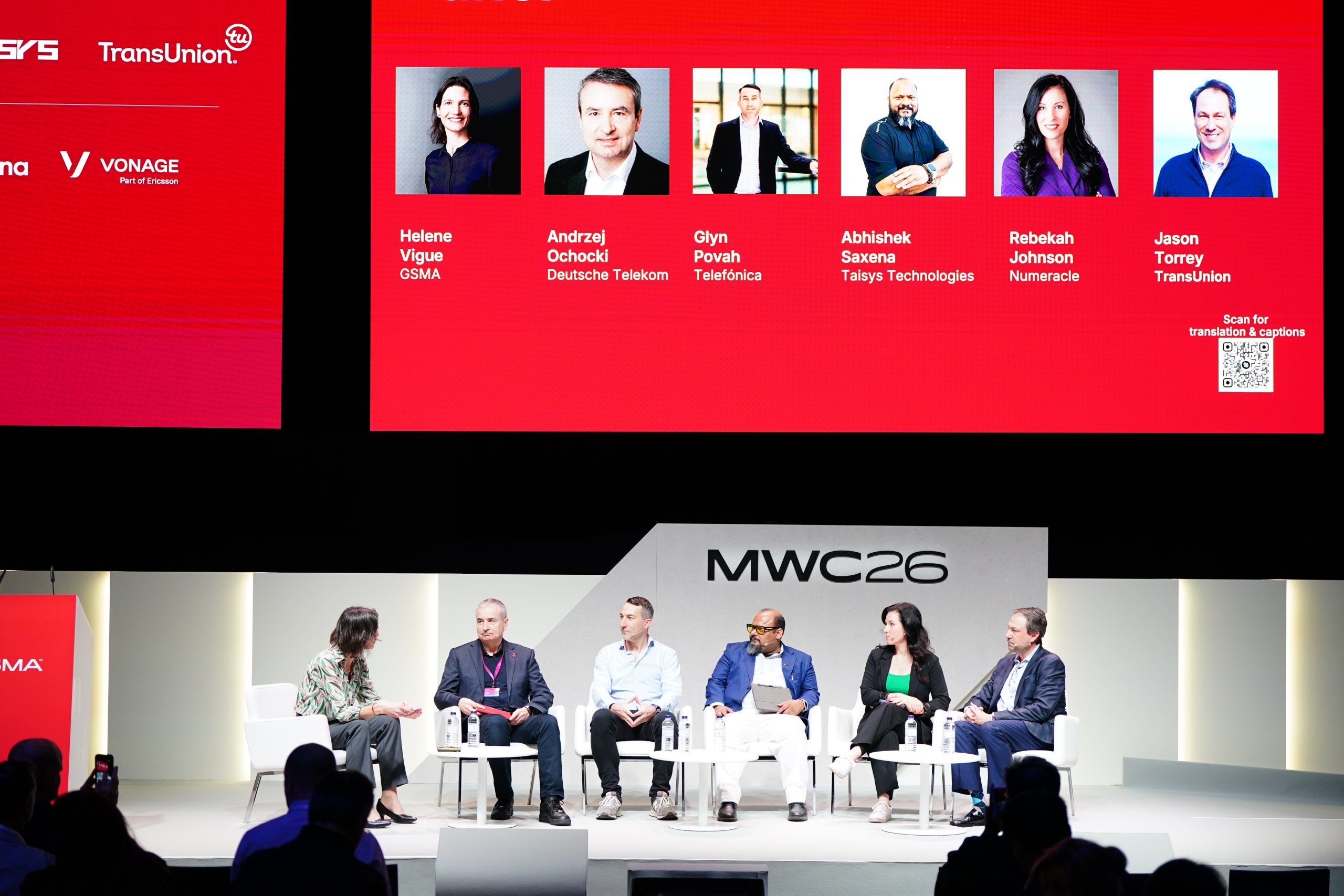 Six people sit on a stage panel at MWC26, with a red screen showing speakers' names and photos behind them.