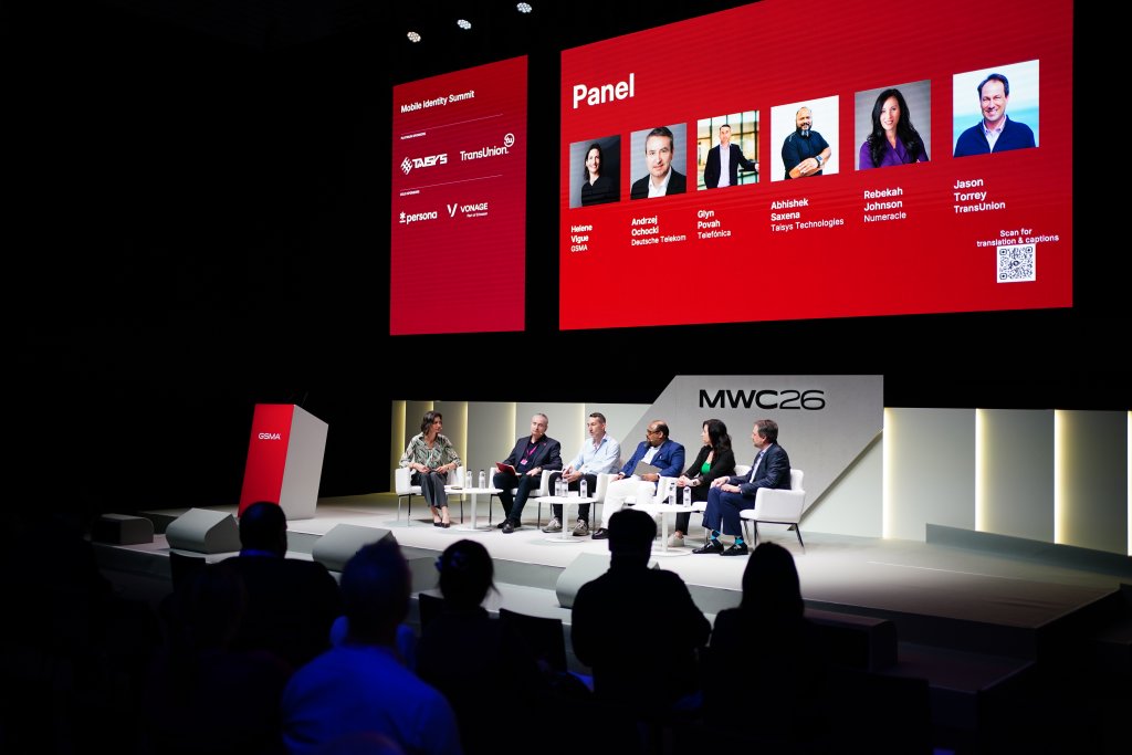 A panel of six people discusses on stage at MWC26, with a red speaker list displayed on a large screen.