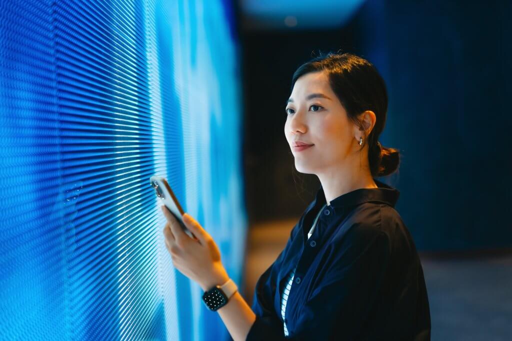 Woman holding a phone stands near a glowing blue LED wall, looking thoughtfully ahead.