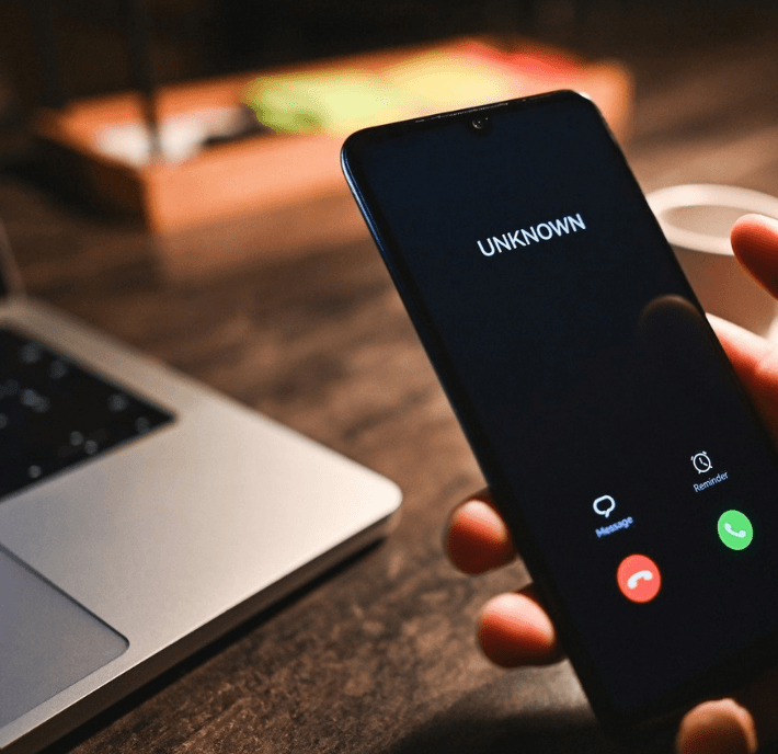 A hand holds a smartphone receiving a call from “UNKNOWN.” The phone screen shows options to answer or decline. A laptop and a cup are on a wooden desk in the background, softly lit by warm lighting.
