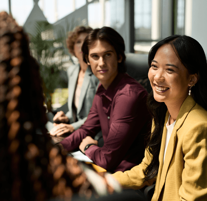 Four people sit at a conference table in a sunlit office, dressed in business attire. A woman in a yellow blazer smiles brightly while a man in a maroon shirt looks on attentively. Others are blurred in the background, engaged in conversation.