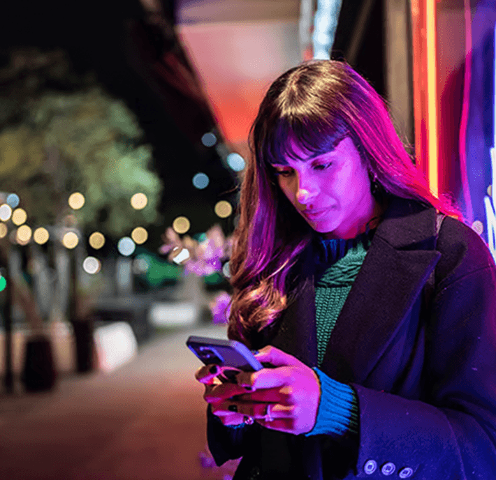 A woman with long dark hair looks down at her smartphone, reflecting on her mobile identity while standing outdoors at night. She wears a coat over a dark sweater, illuminated by bright pink and blue neon lights.