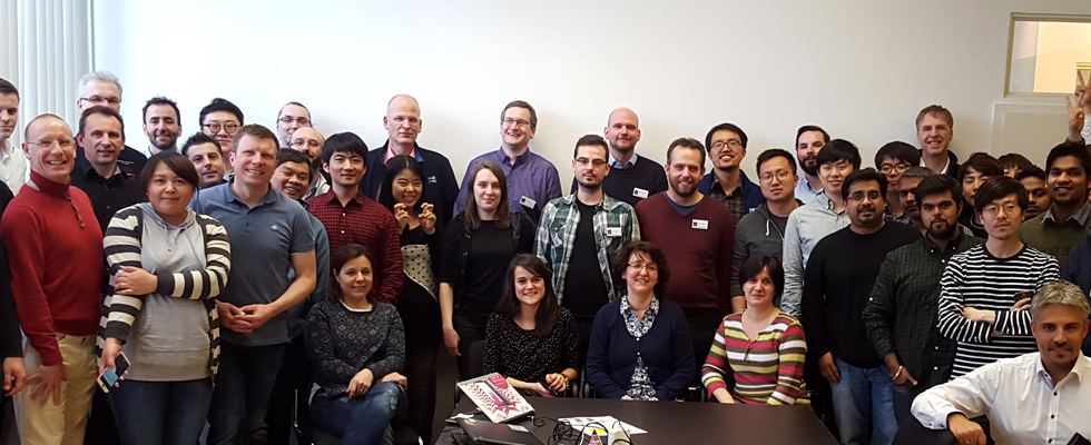 A diverse group of around 35 people, including men and women of various ages, stand closely together in a spacious room. They are dressed casually, some smiling and others with neutral expressions. A table with papers and a laptop is visible in the foreground.