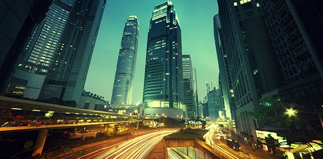 A vibrant cityscape at dusk showcasing tall skyscrapers with glowing lights. The scene includes a long exposure effect, highlighting streaks of car lights on the road below, creating dynamic movement. The sky has a greenish hue, and the buildings stand prominently against it.