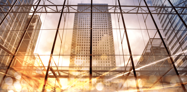 View of a modern cityscape through a large glass window, capturing high-rise buildings under a cloudy sky. The scene is bathed in golden sunlight, creating reflections and bokeh effects on the glass. The structures display a mix of steel and glass architecture.