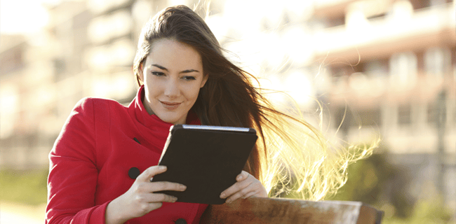 A woman wearing a red coat sits outdoors on a bench, holding a tablet. Her long hair is gently blown by the wind. The background is softly blurred, showing buildings and a sunny sky, suggesting a pleasant day. She appears focused and content while looking at the tablet.
