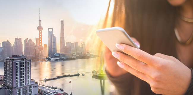 A person holds a smartphone in the foreground. The background features the modern skyline of Shanghai with the iconic Oriental Pearl Tower and other skyscrapers illuminated by a warm sunset. The Huangpu River is visible, reflecting the orange and pink hues of the sky.