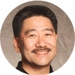 A person with short black hair and a mustache is smiling against a neutral background. They are wearing a black shirt, and their expression is friendly. The lighting is soft, highlighting their facial features.