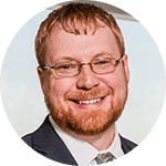 A bearded man with short red hair is smiling. He is wearing glasses and a gray suit with a white shirt and patterned tie. The background is blurred, suggesting an indoor setting.