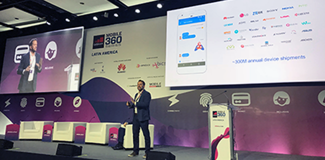 A speaker stands on stage at a tech conference, presenting a slide about mobile device shipments and RCS B2C campaigns, featuring logos of tech companies and a smartphone graphic. The stage backdrop has a purple and white design with the "Mobile 360 Latin America" logo. Conference seating is visible.