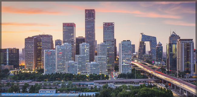 A cityscape at dusk features a vibrant skyline with tall skyscrapers under a colorful sky transitioning from pink and orange to blue. Light trails from busy traffic are visible on a highway below, conveying movement and energy amid the urban landscape.