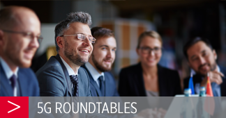 A group of five people in suits are sitting around a table engaged in a discussion or meeting. The focus is on a man with glasses and facial hair, smiling. In the foreground, a banner reads "5G ROUNDTABLES" with a red and gray design. The background is softly blurred.