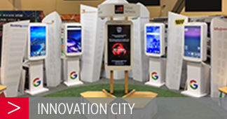 A display at an event named "Innovation City" featuring six digital kiosks. Each kiosk has a digital screen showing various tech images, with Google logos at the base. The central kiosk has a red and black design. The area has a green carpet and wooden platform.