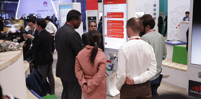 A group of people gathered at an exhibition booth, facing a large screen displaying software. They appear engaged in a discussion. Several attendees wear ID lanyards. Surrounding booths contain electronic displays and branding for various tech companies.