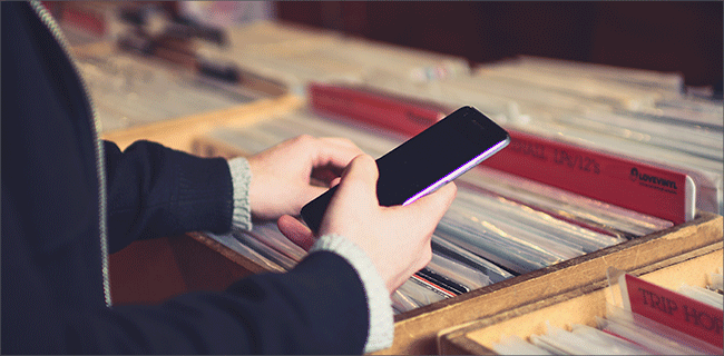 A person, immersed in their smartphone, is browsing through a collection of vinyl records neatly arranged in wooden crates with red dividers. Dressed in a dark coat and sweater, they remain focused on the device's RCS features while exploring the charming analog realm around them.