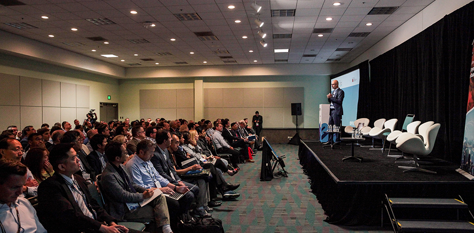 A speaker stands on a stage addressing a large audience in a conference room. The audience is seated in rows, listening attentively. The room has a high ceiling with bright lights, and there is a black curtain backdrop behind the stage. Empty chairs are arranged on the stage next to the speaker.