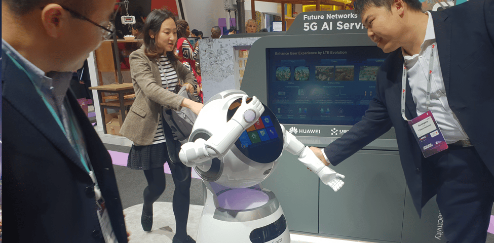 A group of people interacts with a humanoid robot at a tech exhibition. The robot has a screen displaying colorful icons. The backdrop features a sign that mentions "Future Networks" and "5G AI Services." One person extends their hand towards the robot, which is extending its arm back.