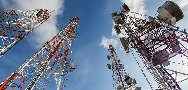 A low-angle view of several telecommunications towers against a blue sky with scattered clouds. The towers are equipped with various antennas and satellite dishes, painted in red and white for safety visibility. The perspective accentuates their towering height and structural complexity.