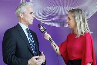 A man in a suit and tie speaks into a microphone held by a woman in a red blouse. They stand in front of a purple backdrop with abstract white patterns. The woman is facing him, seeming engaged in the conversation.