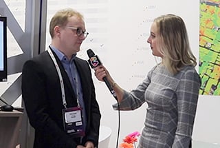 A woman in a gray plaid dress is interviewing a man in a black suit and blue shirt. She holds a microphone with a colorful logo. They stand in a booth with a display screen and colorful diagrams in the background. The man wears glasses and a lanyard with a name badge.