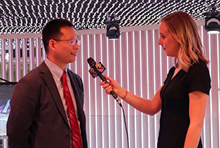 A woman in a black dress interviews a man in a suit and red tie. They stand in a modern room with vertical white slats and a ceiling of small, illuminated bulbs. She holds a microphone with a colorful logo. The atmosphere is professional and well-lit.