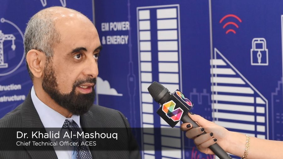 A man in a suit is being interviewed with a microphone held by a person with black nail polish. The backdrop features blue graphics of buildings and icons related to energy. Text overlay reads, "Dr. Khalid Al-Mashouq, Chief Technical Officer, ACES.