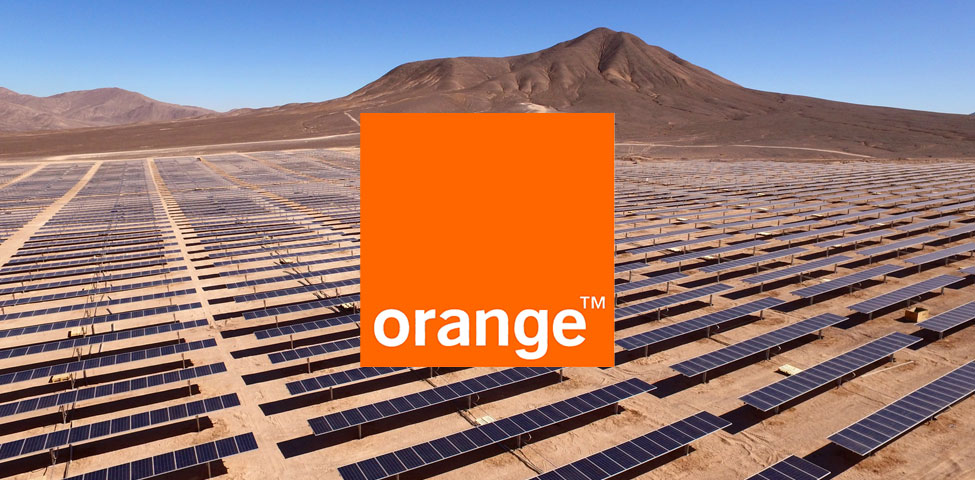 A vast solar farm with rows of solar panels stretches across a desert landscape. In the background, a large brown mountain rises against a clear blue sky. In the center, an orange square with the word "orange" in white text is prominently displayed.