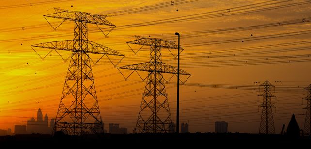 Silhouetted against a vibrant orange sunset, several high-voltage transmission towers dominate the foreground, with numerous lines stretching across the sky. In the background, city skyscrapers are faintly visible, blending into the horizon as the sun sets.