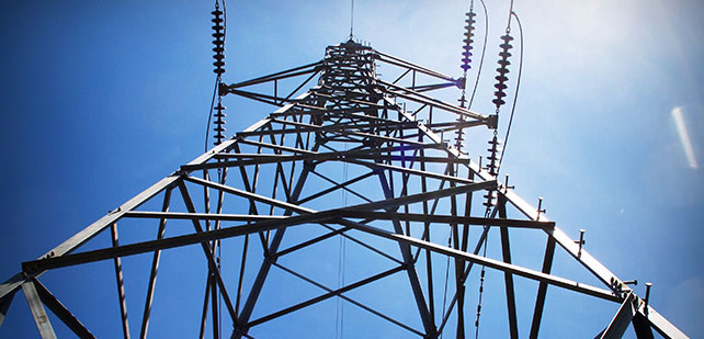 Low-angle view of a metal electricity pylon against a clear blue sky. The sun shines from the top right corner, casting light on the structure. Insulators and cables are visible, and the geometric pattern of the pylon creates a sense of height and strength.